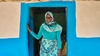 Sekina Hassen standing in a bright blue doorway wearing a striking teal outfit and smiling, Inkomoko 2026