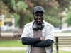 A participant of the North Amercian Mastercard Foundation Alumni Network, standing outdoors in a black baseball hat wearing a light coloured t-shirt with a long sleeved shirt underneath, smiling at the camera.