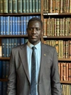 John Bok Chol standing in a smart dark grey suit, standing in front of a large bookshelf.