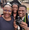 Three people smiling to camera, with the person in the centre in a graduation hat and the the person on the right holding a camera