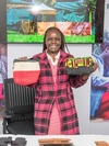 Pauline Akinyi Juma holding some handmade purses, wearing a pink and black plaid sweater over a pink dress with a white collar smiling.