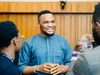 Great Nnamani talking and smiling wearing a light blue button up shirt with a white pattern along the front and pocket 