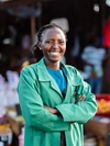 Vegetable trader, Ciru, standing outside at her vegetable stall, wearing a green workcoat and smiling brightly with her arms crossed in front of her chest.