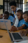 A woman leans over students to help on a laptop.