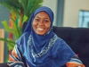 A young woman sitting in an armchair, wearing brightly coloured dress and headscarf smiling.