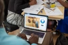 A view over a persons shoulder with them working on a laptop with a desk of papers in front of them