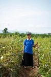 Irene Nimbona standing in a field, holding the long handle of a wooden tool, wearing a long black skirt, bright blue collared t-shirt and a baseball hat.