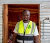 Vasco Hamisi standing outside of a structure in Kakuma refugee camp wearing a high visibility vest with the Okapi Green logo, 2025.