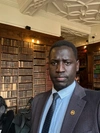 John Bok Chol standing in a smart dark grey suit, standing in front of a large bookshelf.