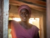 A woman standing in her place of work, wearing a red patterned dress with rose coloured head scarf, Sédhiou, Senegal, 2024.