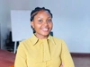 Brenda Mumwesigye smiling and standing with her arms crossed in front of her torso and wearing a mustard yellow button up shirt with a tie collar
