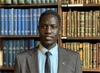 John Bok Chol in a dark grey suit and tie standing in front of a bookcase