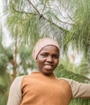Rehema Mgova standing outdoors in front of a tree, smiling, One Acre Farm, March 2026.