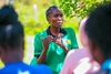 Hyliene Atieno s'adressant à un groupe en plein air, portant un t-shirt vert vif avec un col noir et les marques de la Mastercard Foundation et de Practical Action