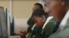Young person in a classroom, in front of a computer wearing a dark green uniform with a white collared shirt