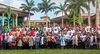 Large group of people stand together facing the camera with buildings a palm trees in the background