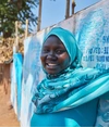 Sekina Hassen, standing outdoors in front of some street artwork, wearing a striking teal shirt and head covering, supported by partner Inkomoko 2026