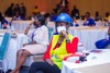 Nyabonyo Anna Charity sitting at a table in a conference room in a high-visibility vest, blue hard hat and a medical face mask