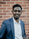 Hamdi Mohamed smiling in a smart dark grey suit jacket and white button up collared shirt.