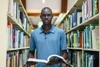 Man in blue polo shirt stands in a library aisle holding an open book looking towards the camera 