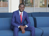 Smiling man in a smart blue suit with a red tie sat on a blue sofa 