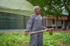 Monica Jackson standing outdoors at her farm, wearing grey coveralls holding a farming tool and smiling, in Kakuma refugee camp, Kenya, 2026.