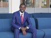 Amos Kwizera smiling to camera in a smart blue suit sat on a blue sofa