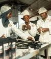 Three young people working in a food manufacturing factory, wearing protective clothing and head coverings.