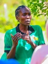 Hyliene Atieno s'adressant à un groupe en plein air, portant un t-shirt vert vif avec un col noir et les marques de la Mastercard Foundation et de Practical Action