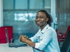 A woman sits at a desk with hands folded above her laptop smiling at the camera, Nancy Telecel, Ghana, 2024.