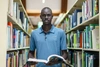 Jacob stands in a library looking to camera holding open a book in front of him