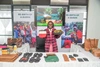Pauline Akinyi Juma tenant des sacs à main sur un stand d'articles en cuir fabriqués à la main, portant un pull à carreaux rose et noir sur une robe rose à col blanc, souriante.
