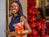 A young woman standing outdoors holding a large bag of fresh tomatoes holding a cell phone, Big Samps, Ghana, 2024.