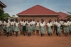 A group of schoolchildren in front of a brick building, smiling and walking toward the camera.