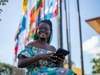 A Mastercard Foundation Scholar at EARTH U, sitting outdoors holding a tablet, smiling confidently, with a number of colourful flags behind her.