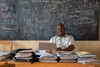 A teacher sitting at their desk at the front of the class in front of a blackboard with chalk writing of mathematical formulas