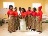 Sarah Kachedwa standing in a storage room with large bags of grains, with a group of women workers wearing similar clothes, red tshirt and patterned skirts, as part of the YEFFA program in partnership with AGRA.