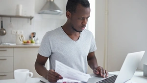 Young man sitting at a table using a laptop. He's holding a bill and has a red calculator next to him.