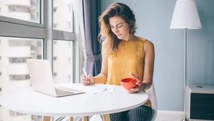 A woman in a yellow sleeveless top sits at a table by a window writing on paper. She holds an orange teacup and has a laptop open in front of her. There's a floor lamp in the background.