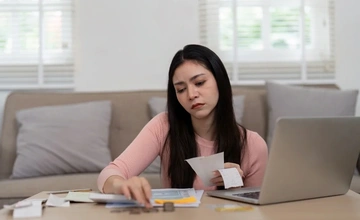 young woman sat at her dining table looking through documents and receipts with her laptop open