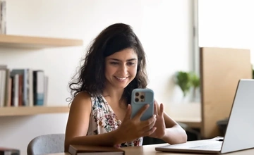 A woman sat at her desk is checking her credit score on her mobile phone and smiling