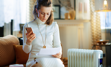 Woman wrapped up next to a radiator paying her energy bills on her mobile.