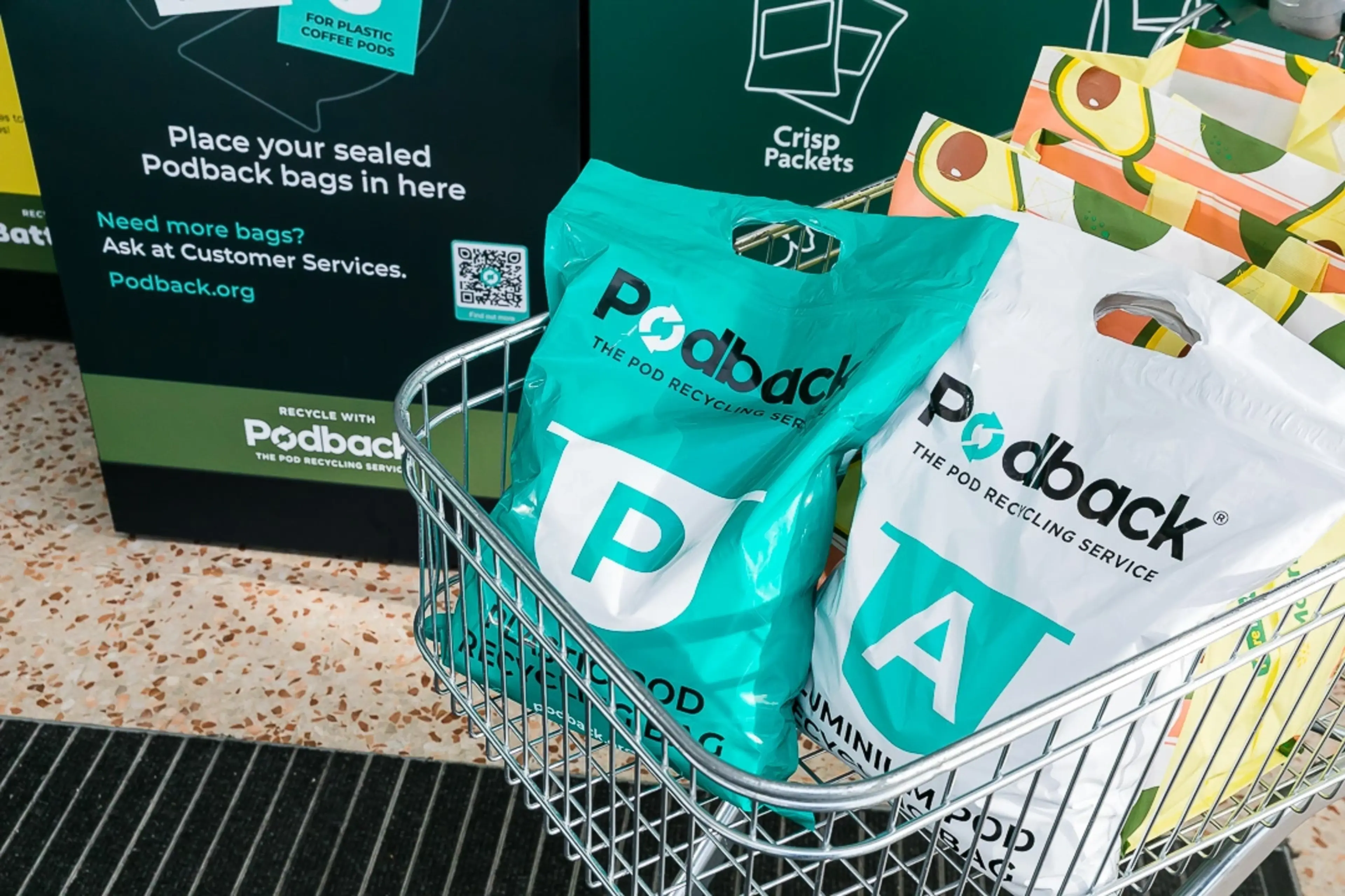 A shopping trolley with green and white Podback bags next to a recycling station in a supermarket