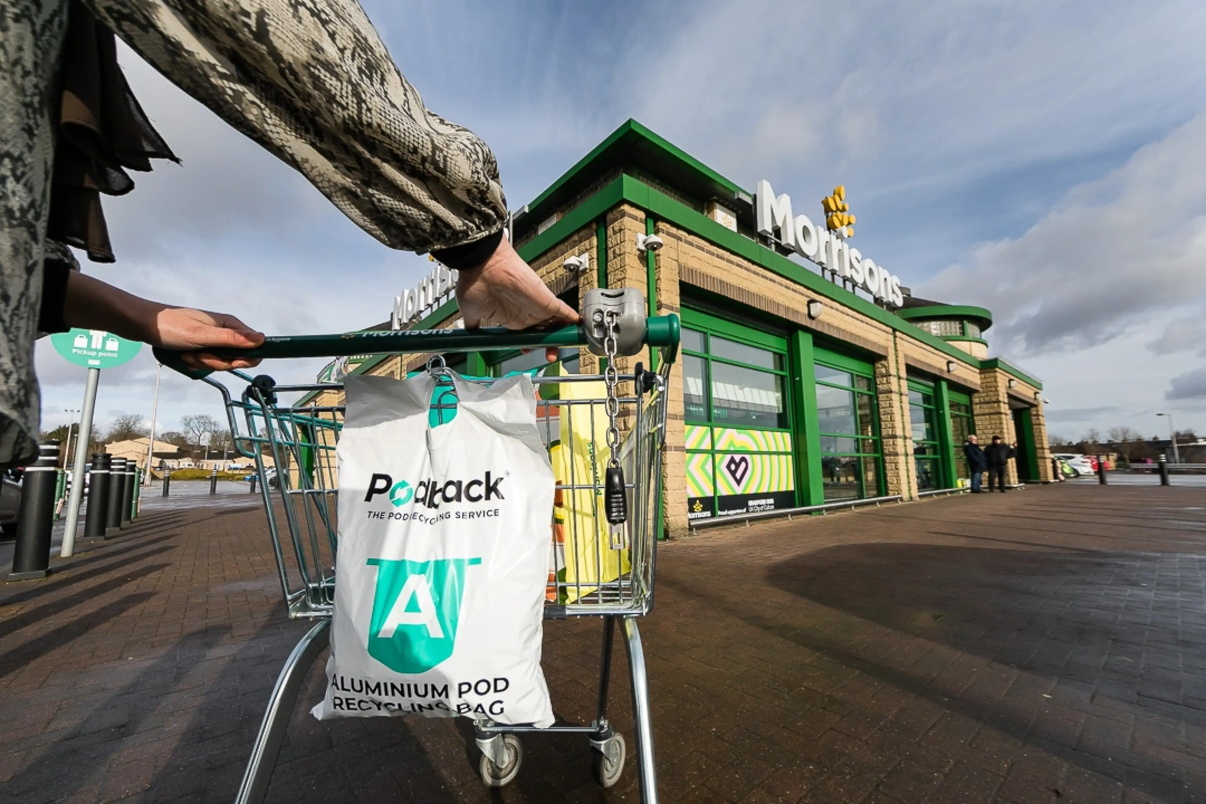 A person pushing a shopping trolley containing a Podback recycling bag towards a Morrisons supermarket