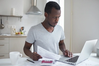 Young man sitting at a table using a laptop. He's holding a bill and has a red calculator next to him.