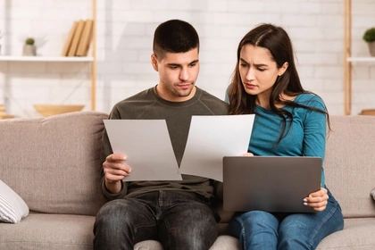 Young couple sit on a grey sofa with a laptop and bills looking concerned