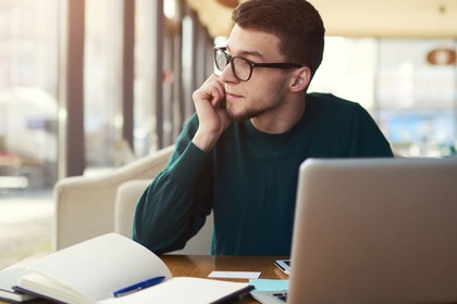 Man in glasses looking out of the window with a notebook and a laptop on the table in front of him