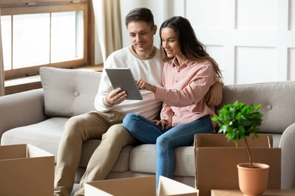 happy couple sat on sofa looking at their tablet, surrounded by cardboard boxes