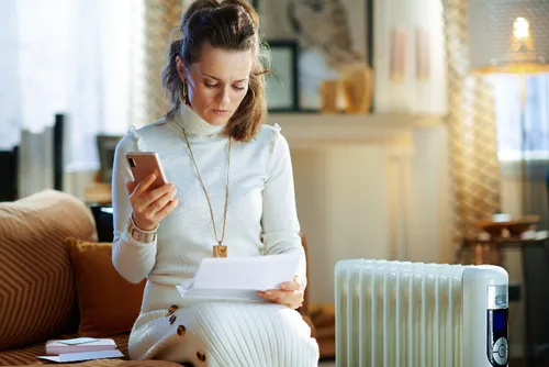 Woman wrapped up next to a radiator paying her energy bills on her mobile.