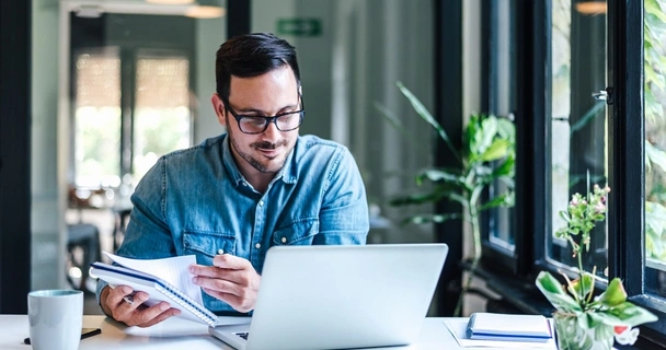 man looking at his computer while holding a notebook